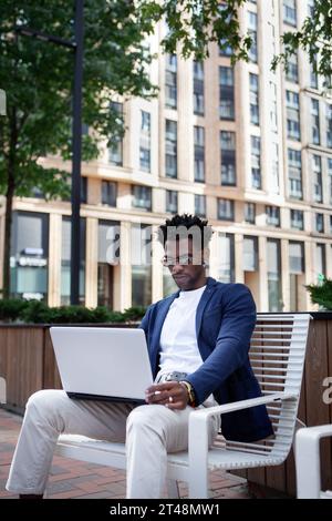 Afroamerikaner mit Laptop arbeitet in der Stadt. Ernster männlicher Freelancer sitzt im Anzug mit Jacke. Stockfoto