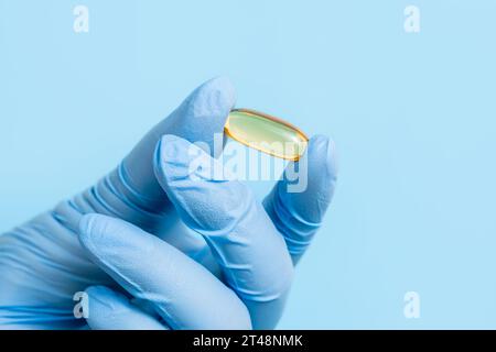 Arzt, Apotheker oder Wissenschaftler Hand in blauen Handschuhen mit Omega-3-Kapsel. Vitaminpille oder -Ergänzung auf blauem Hintergrund. Gesundheitswesen und Wissenschaft Stockfoto