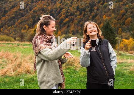 Zwei lachende Frauen mit Glühwein auf herbstlichem Naturhintergrund. Zwei Schwestern haben Spaß. Fröhliche Mädchen in den Herbstbergen. Glückliche Freunde im Park. Stockfoto