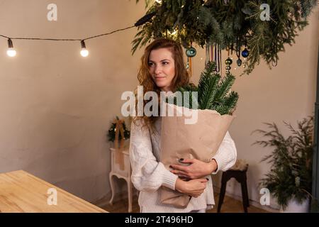 Porträt einer jungen Frau in weißem Strickpullover, mit umweltfreundlichem Winterstrauß in der Hand, Fichtenzweigen in Bastelpapier. Authentisches Geschenk für Urlaub Stockfoto