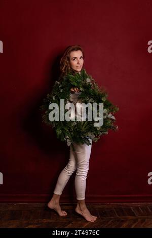 Glückliche, stilvolle junge Frau im gestrickten weißen Pullover hält grünen Weihnachtskranz in den Händen auf dunklem burgunderroten isolierten Hintergrund. Platz für Text. Millen Stockfoto