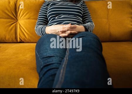 Eine nicht erkennbare Frau in einem gestreiften Pullover und einer blauen Jeans sitzt mit nach vorne gestreckten Beinen und gekreuzten Daumen auf dem Sofa. Das Mädchen ruht sich aus Stockfoto