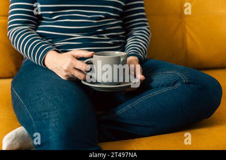 Eine nicht erkennbare Frau in einem gestreiften Pullover sitzt auf dem Sofa und hält eine Tasse Kaffee in der Hand. Unwiedererkennbare glückliche Mädchen trinkt heiße köstliche t Stockfoto