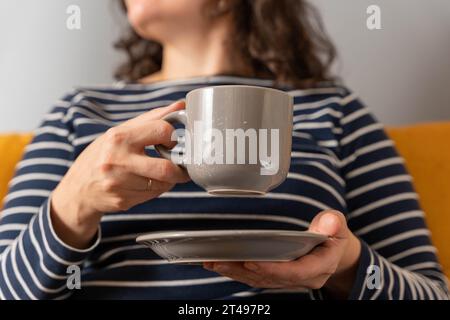 Eine nicht erkennbare Frau in einem gestreiften Pullover sitzt auf dem Sofa und hält eine Tasse Kaffee in der Hand. Unwiedererkennbare glückliche Mädchen trinkt heiße köstliche t Stockfoto