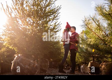 Fröhliches verliebtes Paar in karierten roten Hemden, Strickmützen täuschen herum und lachen auf dem grünen Weihnachtsbaummarkt. Der junge Mann hält die Hände Stockfoto