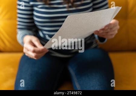 Eine nicht erkennbare Frau sitzt auf einem gelben Sofa und hält ein leeres Blatt Papier in der Hand. Ihre Anzeige. Frau zeigt Poster, Leinwand, weißes Blatt Papier Stockfoto