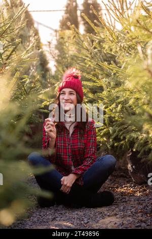 Junge Frau mit Glitzer in der Hand, mit rotem kariertem Hemd, gestricktem Hut unter grünem Weihnachtsbaummarkt. Lockiges Mädchen lacht und freut sich Stockfoto