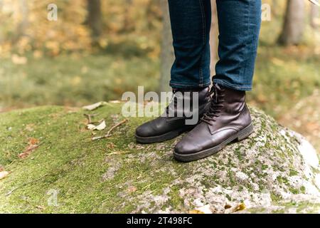 Damenfüße in herbstlichen Lederstiefeln und Jeans stehen auf einem großen Felsbrocken, der mit grünem Waldmoos bedeckt ist. Spaziergänge im Wald. Natur und aktives Rezidiv Stockfoto