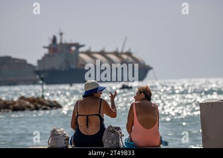 Zwei Damen in Badeanzügen genießen den Strand mit der Armada LNG Mediterrana in der Ferne, einer schwimmenden Gasspeicher- und -Rückvergasungseinheit in Malta. Stockfoto