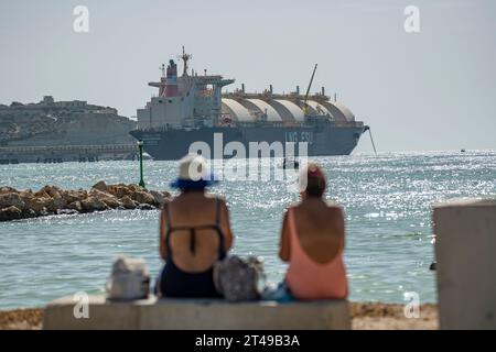 Zwei Damen in Badeanzügen genießen den Strand mit der Armada LNG Mediterrana in der Ferne, einer schwimmenden Gasspeicher- und -Rückvergasungseinheit in Malta. Stockfoto