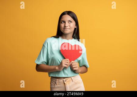 Niedliches brünettes Teenager-Mädchen, das rotes Papierherz in den Händen hält und auf gelbem Hintergrund posiert. Stockfoto