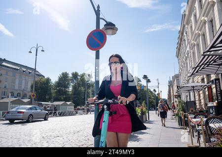 Im Sommer im Zentrum von Helsinki, Finnland, saust eine Frau in Sommerkleid und Mantel auf einem Elektroroller über den Bürgersteig. Foto: Rob Watkins Stockfoto