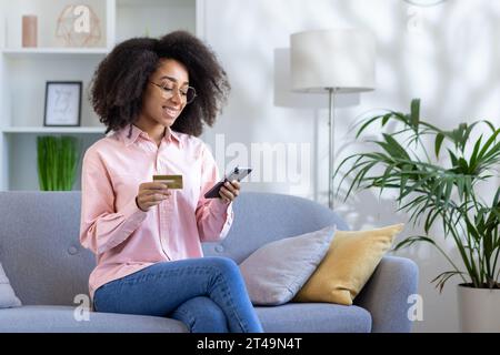 Junge schöne Frau sitzt auf Sofa im Wohnzimmer zu Hause, afroamerikanische Frau lächelt zufrieden mit App auf dem Telefon, mit Bankkreditkarte für Online-Shopping. Stockfoto
