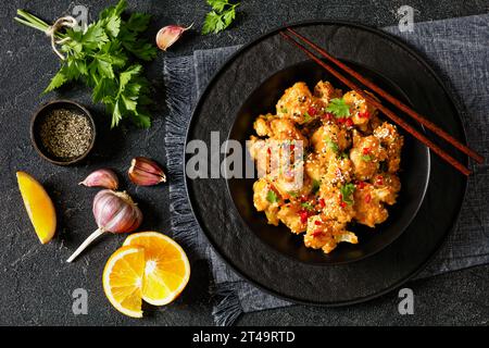 Knusprige Blumenkohlblüten mit Sesamsamen, Chiliflocken und Petersilie in schwarzer Schüssel auf Betontisch mit Zutaten und Stäbchen Stockfoto