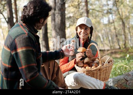 Glückliche junge Frau mit einem Korb voller Pilze, die ihren Mann mit frischem Boletus ansieht, während sie eine Pause im Wald genießt Stockfoto