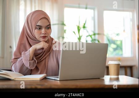 Eine konzentrierte junge asiatisch-muslimische Frau, die einen Hijab trägt, schaut mit einem ernsthaften, nachdenklichen Gesicht auf ihren Laptop-Bildschirm, plant oder denkt etwas Stockfoto