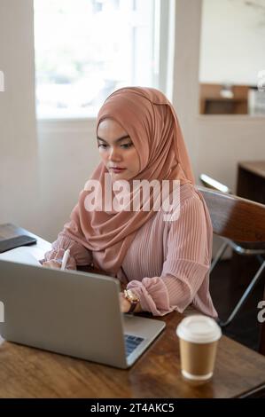 Eine konzentrierte und nachdenkliche asiatisch-muslimische Frau arbeitet an ihrem Laptop, während sie in einem Café sitzt. Geschäftsfrau, Freiberuflerin, Studentin Stockfoto