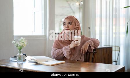Eine nachdenkliche asiatisch-muslimische Frau, die einen Hijab trägt, schaut von der Kamera weg und denkt nachdenklich etwas nach, während sie ihren Morgenkaffee in einer Kiste trinkt Stockfoto