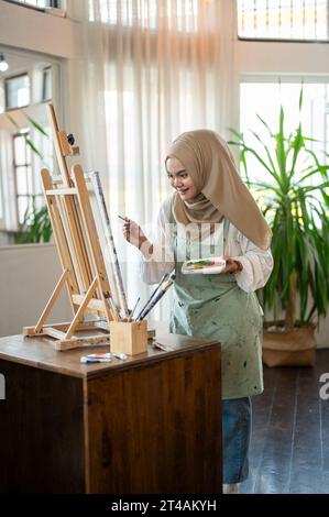 Eine fröhliche und kreative asiatisch-muslimische Künstlerin in einem Hijab und einer Schürze malt in ihrem Atelier auf Leinwand. Eine muslimische Frau, die sich einer Kunstwerkstatt anschließt, Draw Stockfoto