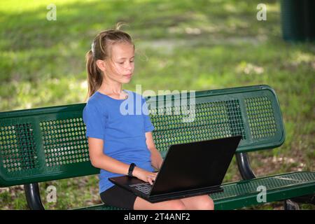 Nettes Teenager-Mädchen tippen auf Laptop-Computer sitzen auf der Bank im Freien am sonnigen Sommertag. Bildung während Quarantänekonzept Stockfoto