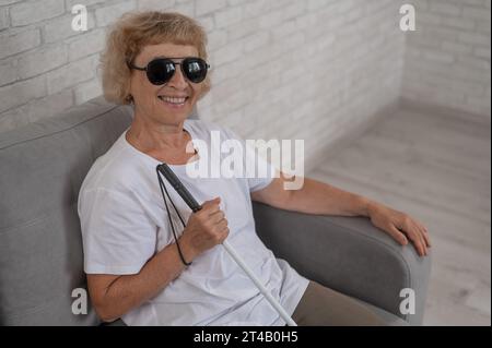 Eine ältere blinde Frau mit Sonnenbrille und einem tastbaren Stock sitzt auf dem Sofa. Stockfoto