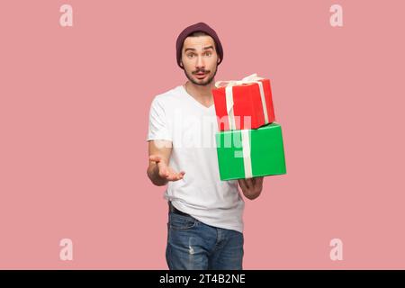 Porträt eines verwirrten bärtigen Mannes in weißem T-Shirt und Beany-Hut, der zwei Geschenkboxen in der Hand hält und mit überraschtem Ausdruck in die Kamera blickt. Innenstudio, isoliert auf rosa Hintergrund Stockfoto
