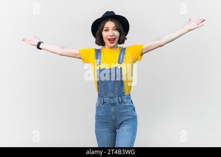 Porträt der staunend aufgeregten, glücklichen Hipster-Frau in blauem Denim-Overall, gelbem T-Shirt und schwarzem Hut, spreizt ihre Arme, zeigt willkommene Geste, einladend. Innenstudio isoliert auf grauem Hintergrund Stockfoto