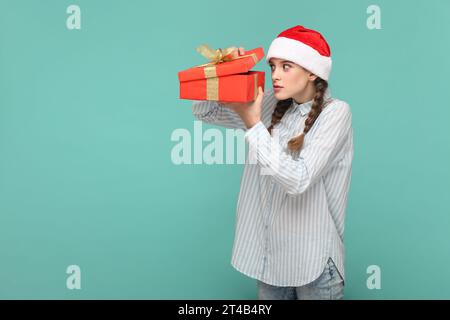 Porträt eines verängstigten Teenagers mit Zöpfen, gestreiftem Hemd und Weihnachtsmann-Hut, Geschenkbox in der Hand und mit großen Augen hinein schauen. Innenstudio, isoliert auf grünem Hintergrund. Stockfoto