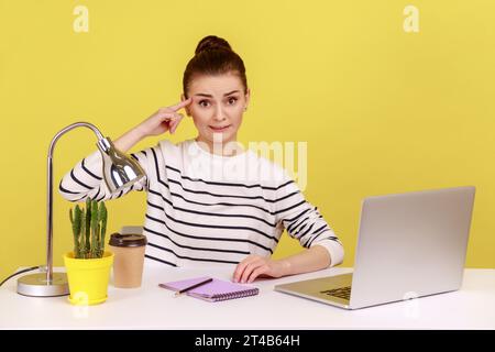 Büroangestellte zeigt dumme Geste mit dem Finger, der mit unzufriedenem müden Ausdruck in die Kamera blickt, am Arbeitsplatz mit Laptop sitzt. Studio im Innenbereich, isoliert auf gelbem Hintergrund. Stockfoto