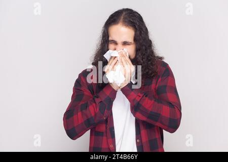 Mann mit langen Haaren im karierten roten Hemd niest im weißen Gewebe, leidet an Rhinitis und laufender Nase, hat Allergie auf etwas, sieht ungesund aus. Innenstudio isoliert auf grauem Hintergrund. Stockfoto