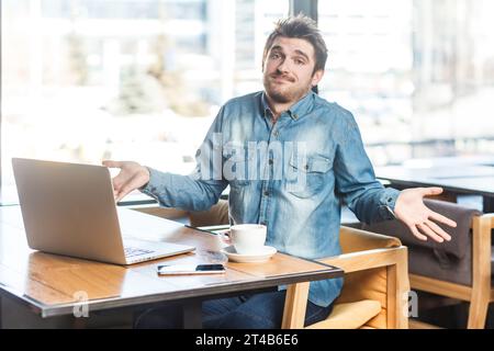 Porträt eines unsicheren jungen Mannes in blauem Hemd, der an einem Laptop arbeitet, Schultern zuckt, nicht weiß, was er beantworten soll, in die Kamera schaut. Innenaufnahme in der Nähe eines großen Fensters, Café-Hintergrund. Stockfoto