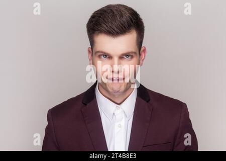 Porträt eines verrückten Comics, ein toller junger Mann, der mit lustigen Augen in die Kamera blickt, in violettem Anzug und weißem Hemd. Innenstudio isoliert auf grauem Hintergrund. Stockfoto