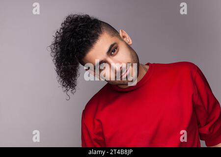 Porträt eines gutaussehenden, lächelnden bärtigen Mannes mit lockigem Haar, neigt seinen Kopf zur Seite, blickt ruhig in die Kamera, trägt roten Pullover. Innenstudio isoliert auf grauem Hintergrund. Stockfoto