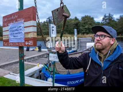 Stolpe, Deutschland. Oktober 2023. Ein Mann schlägt am Anleger der kleinen Passagierfähre über die Peene auf ein Eisen und informiert den Fährmann über das Signal. Von März bis Oktober können Wanderer oder Radfahrer die fast 100 Meter über den Fluss überqueren. Die nun zu Ende gehende Saison könnte jedoch ihre letzte sein – die einzige Fußgängerfähre zwischen Anklam und Jarmen ist wegen fehlender finanzieller Unterstützung durch den Bezirk von der Schließung bedroht. Die Gemeinde hat daher eine Kampagne zur Rettung der Fähre gestartet. Jetzt werden 2.000 Unterschriften gesammelt, bis Credit: dpa/Alamy Live News Stockfoto