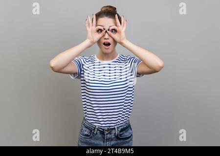 Porträt einer Frau, die ein gestreiftes T-Shirt trägt und durch ein Fernglas blickt und Überraschung ausdrückt, zoomt und Distanz erforscht. Innenstudio isoliert auf grauem Hintergrund. Stockfoto