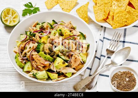 Thunfischsalat mit Avocado, roten Zwiebeln, Gurke und Kapern mit Olivenöl-Limonen-Petersilie-Dressing in weißer Schüssel auf weißem Holztisch, horizontale Sicht von Stockfoto