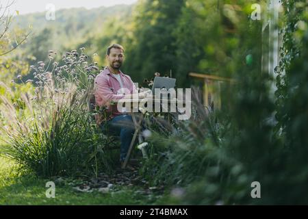 Mann, der im Garten arbeitet, mit Laptop auf den Beinen. Geschäftsmann, der aus der Ferne vom Heimbüro im Freien arbeitet und über neue Geschäfte oder kreative Ideen nachdenkt. Stockfoto