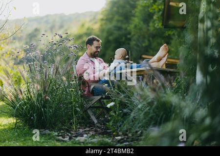 Mann sitzt im Garten mit Füßen auf dem Tisch und spielt mit seinem kleinen Sohn. Vater hat Bindungsmoment mit seinem kleinen Kind. Stockfoto