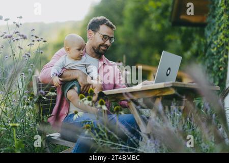 Vater hält Baby in der Hand, während er im Garten am Laptop arbeitet. Geschäftsmann, der aus der Ferne vom Home Office im Freien arbeitet und sich um den kleinen Sohn kümmert. Lebensarbeit Stockfoto