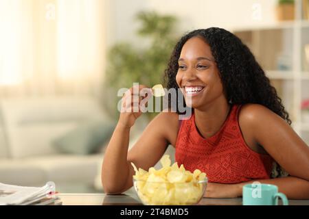 Glückliche schwarze Frau, die Kartoffelchips isst und nach Hause wegschaut Stockfoto