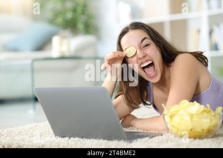 Glückliche Frau mit Laptop, die mit Kartoffelchips spielt, die zu Hause auf dem Boden liegen Stockfoto