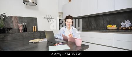 Das Porträt einer brünetten Frau macht Hausaufgaben, arbeitet von zu Hause aus, bereitet sich auf die Untersuchung in der Küche vor, sitzt mit Notebook und Laptop Stockfoto