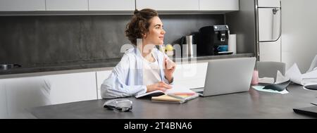 Porträt einer jungen Frau, die denkt, zu Hause an einem Laptop arbeitet, sich Notizen macht, während sie ihre Hausaufgaben macht, Online-Kurse studiert, zuhört Stockfoto