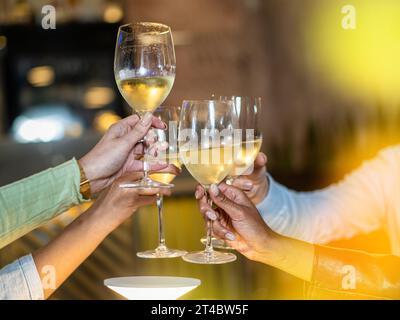 Eine Nahaufnahme von Freunden, die ihre Gläser zum Toast hochheben und mit gekühltem Weißwein feiern. Der goldene Farbton des Ambientes unterstreicht die Wärme von Stockfoto