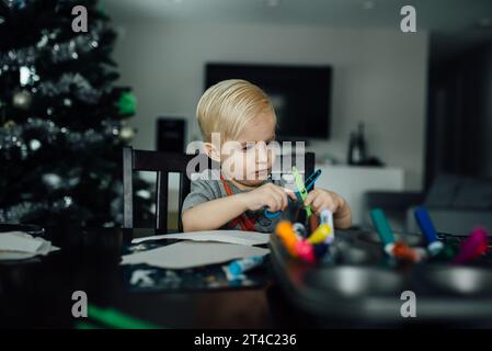 Der kleine Junge schaut sich eine Schere an, während er an einem Tisch bastelt Stockfoto