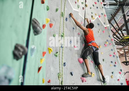 Gutaussehender afroamerikaner in orangefarbenem Hemd, der an Felsbrocken greift und aufblickt, Sportsman Stockfoto