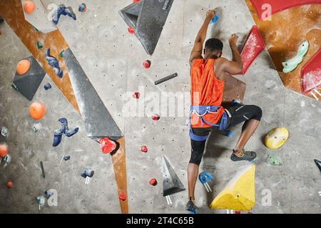 Junger afroamerikanischer Mann mit alpinem Gurtzeug, der die Kletterwand hinaufsteigt, Rückansicht, Bouldern Stockfoto