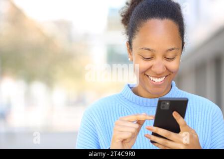 Vorderansicht Porträt einer glücklichen schwarzen Frau mit Handy auf der Straße mit Kopierraum Stockfoto