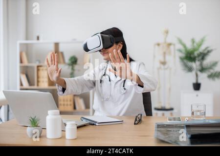 Indische Krankenschwester, die Laborkittel mit VR-Brille trägt, während sie mit modernen Geräten am Schreibtisch sitzt, auf verschwommenem Hintergrund des Schranks. Therapeutin in der Vorbereitung Stockfoto