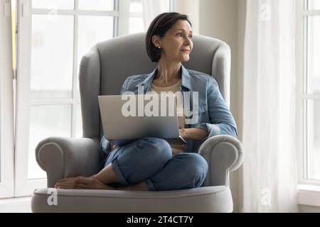 Eine ruhige, reife Frau ließ sich auf einem bequemen Sessel mit Laptop nieder Stockfoto